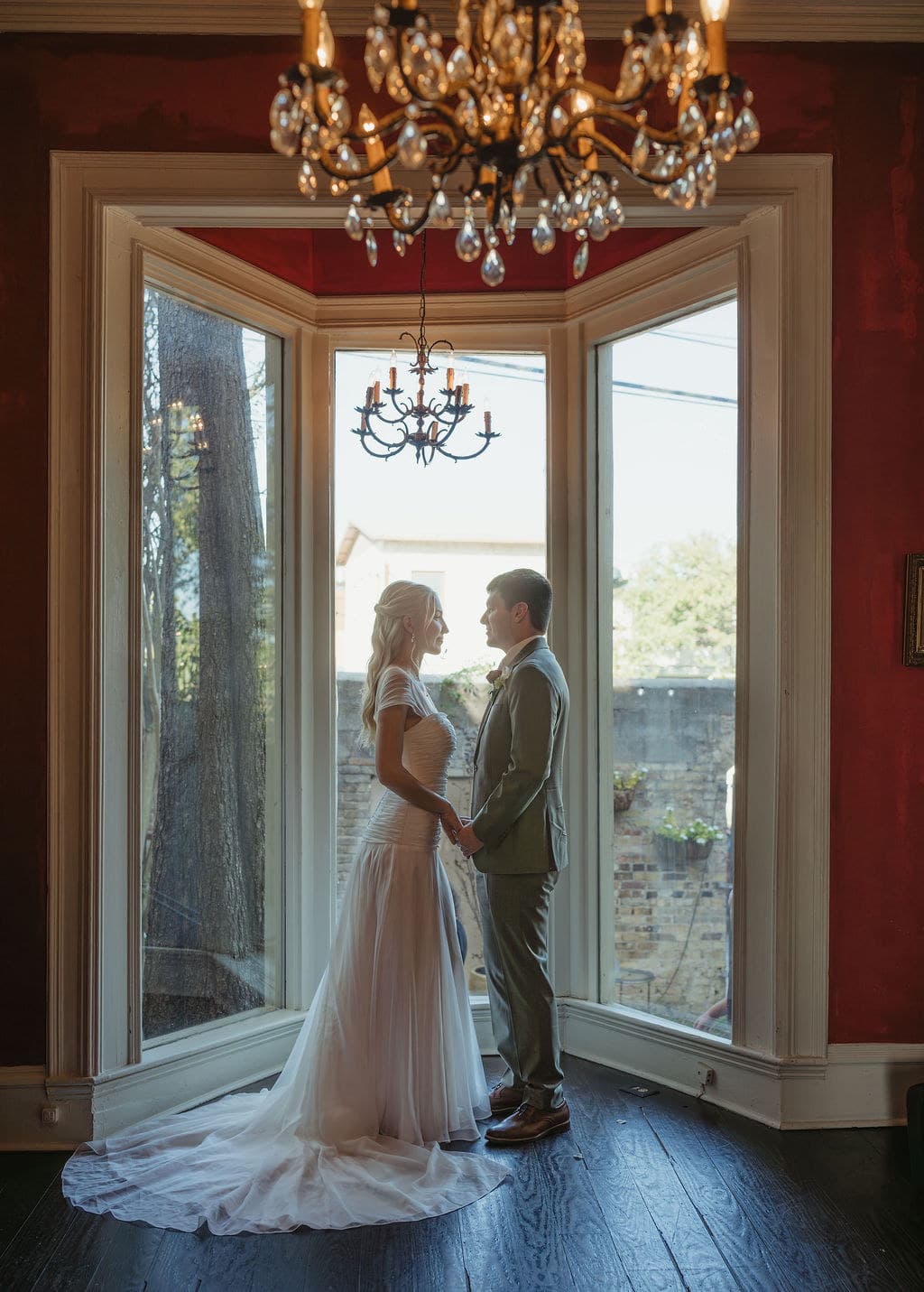 Ben and Elizabeth hold hands in a bay window beneath crystal chandeliers.