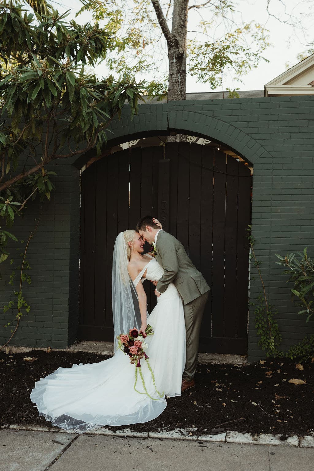 Ben and Elizabeth lean together beneath a green brick arch, with her train spread beside them.